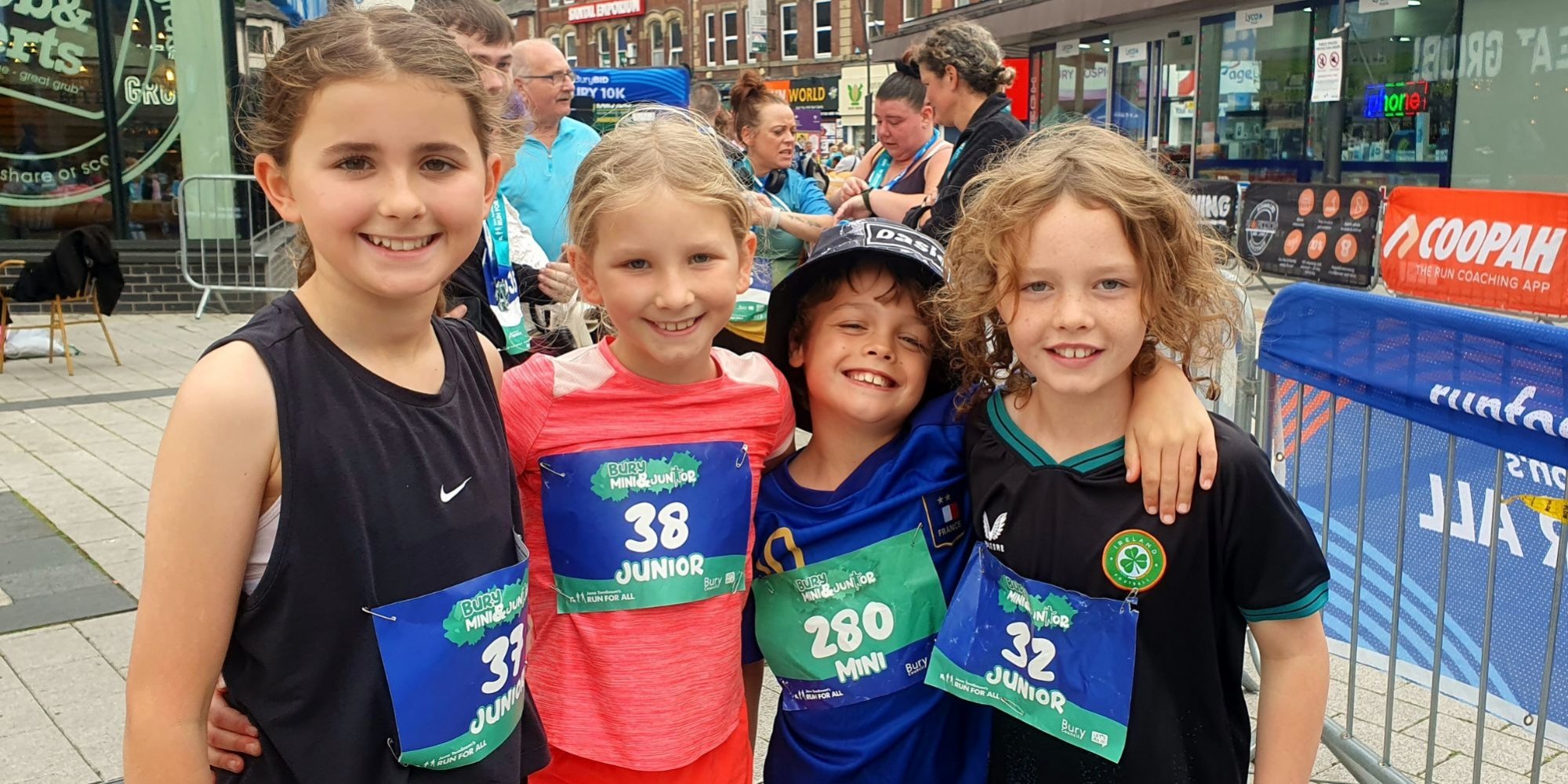 Photo of 4 children with their arms round each other and wearing runner entry numbers