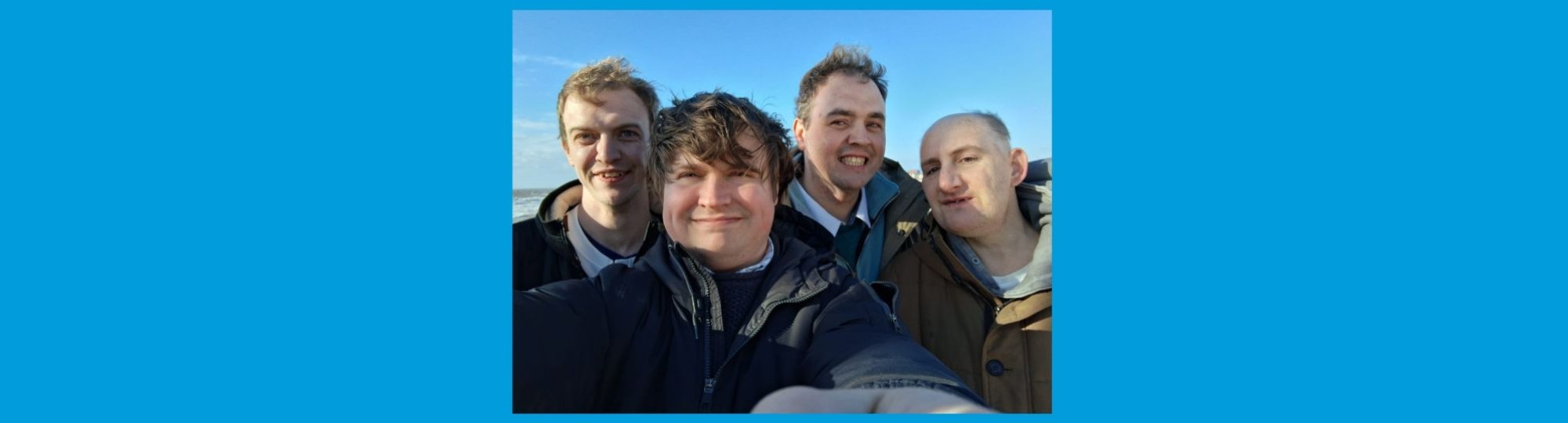 photo: close up of four men smiling to camera. It's sunny and the wind is blowing.
