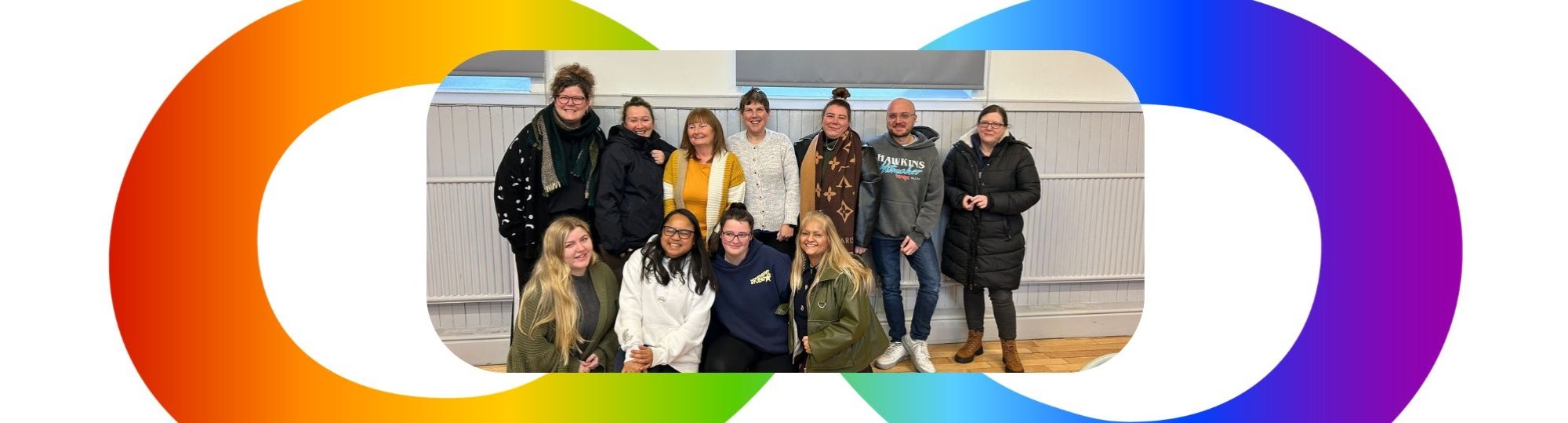 photo of staff members smiling to camera on a training day. Background shows a rainbow coloured infinity symbol