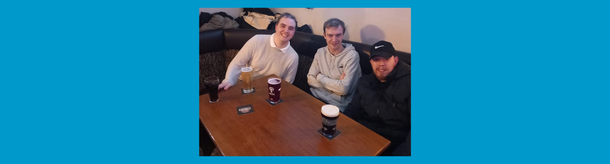 photo: three men leaning together, smiling to camera. They are at a pub table, each with a pint in front of them