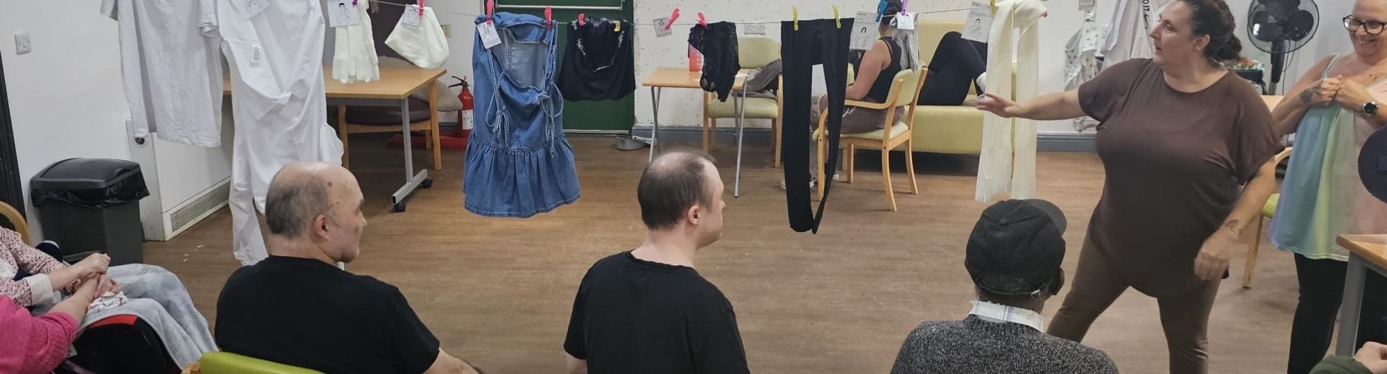 photo: men and women sitting, facing a woman who's standing and signing next to some laundry on a washing line